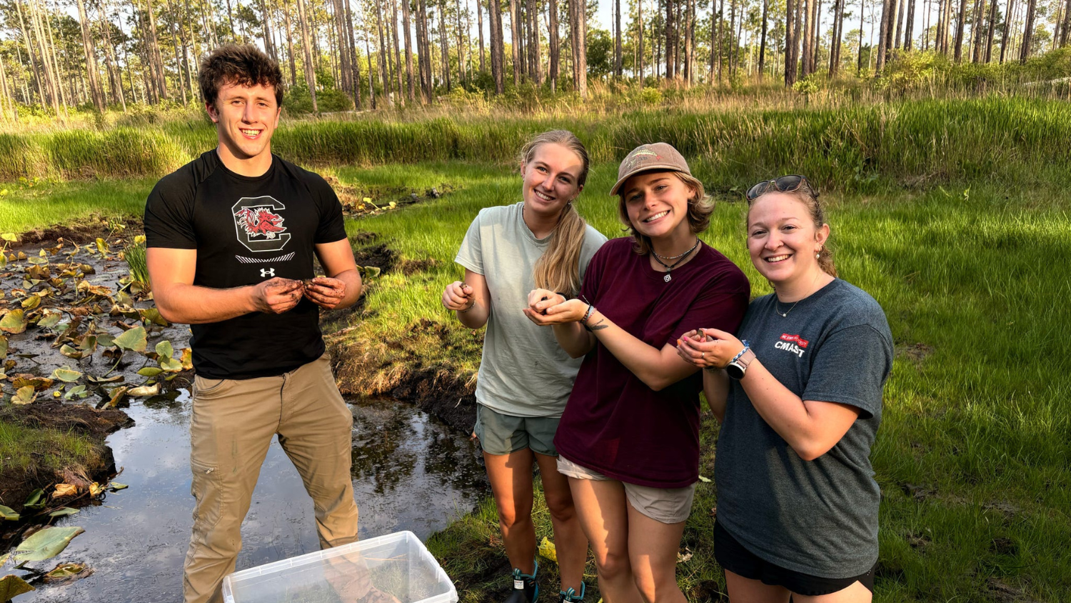 Four people stand in a wooded pond area holding frogs in their hands